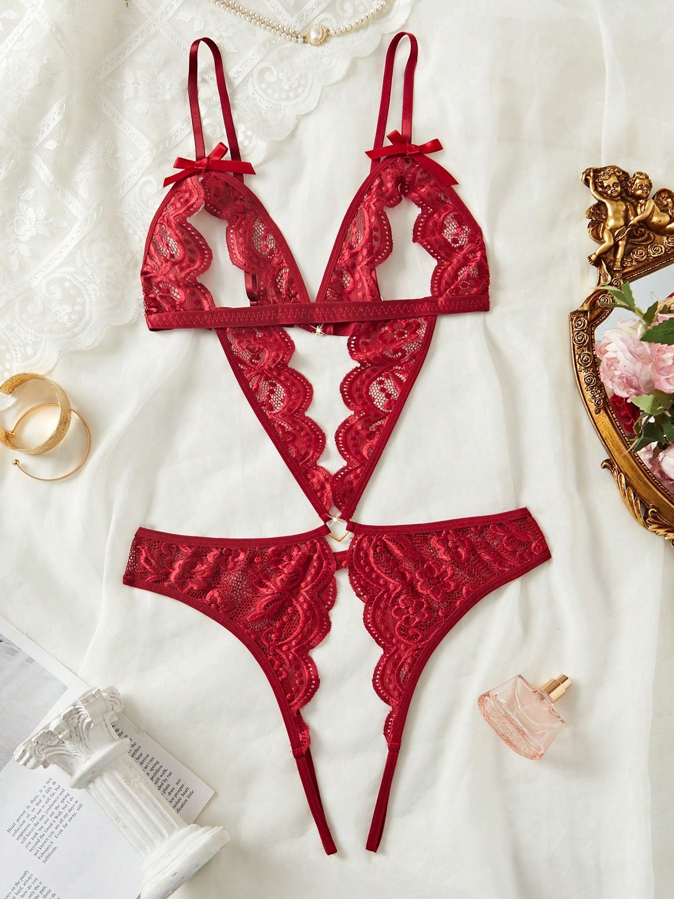 Women's burgundy teddy lingerie bodysuit featuring adjustable spaghetti straps, bow decor, exposed chest design, scalloped trim, metal heart detail, backless semi-sheer contrast lace, and thong-style low-rise bottom.