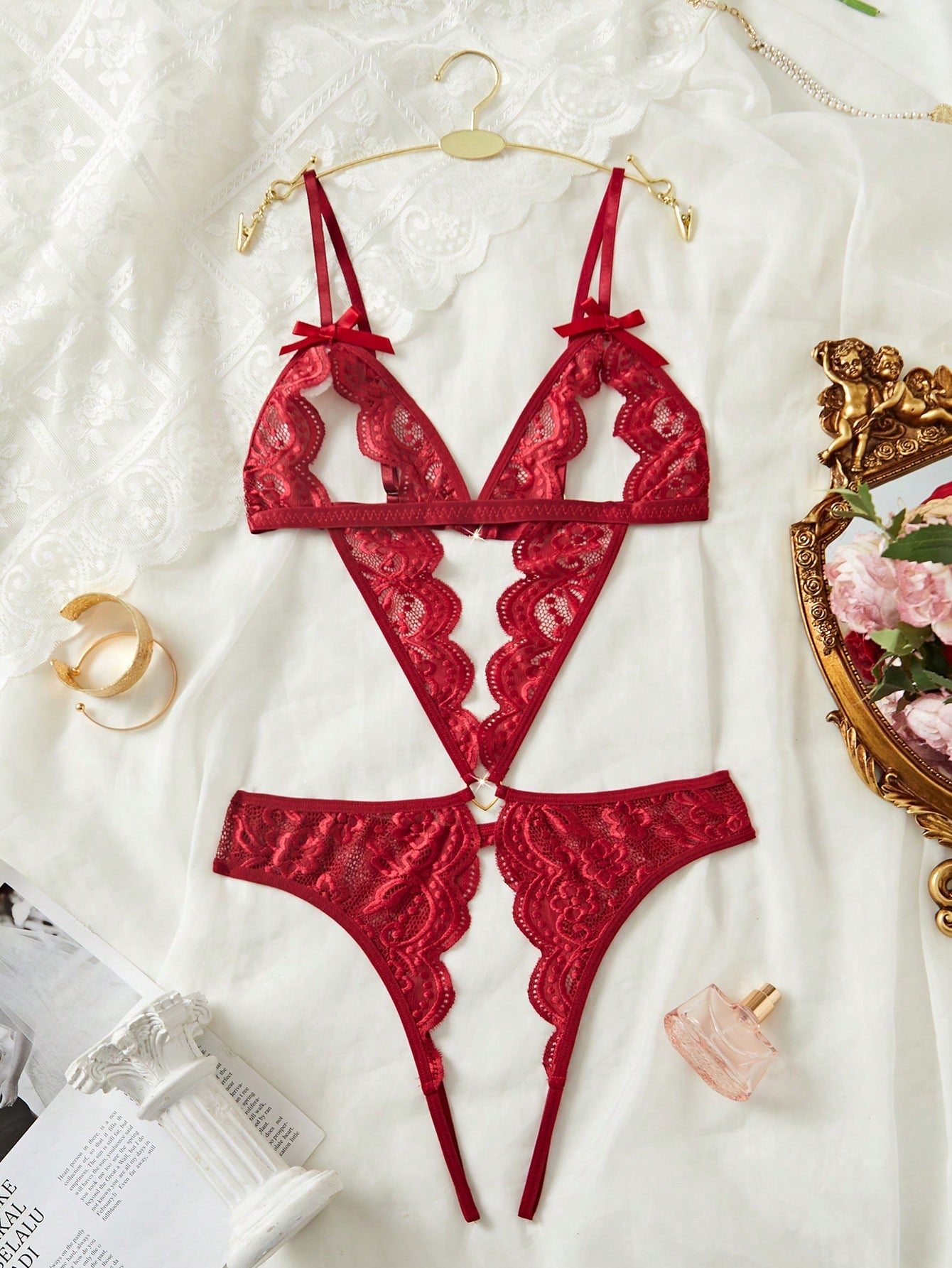 Women's burgundy teddy lingerie bodysuit featuring adjustable spaghetti straps, bow decor, exposed chest design, scalloped trim, metal heart detail, backless semi-sheer contrast lace, and thong-style low-rise bottom.